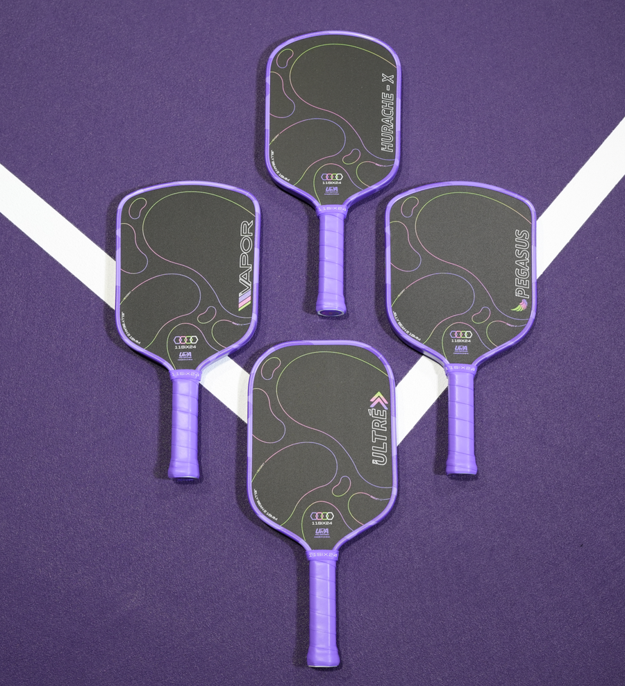 Four pickleball paddles arranged on a purple court surface with white boundary lines. The paddles have purple handles and borders, dark gray paddle faces with abstract curved line graphics, and visible Audi branding.